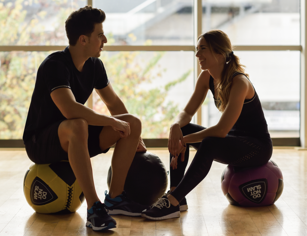 Deux amis en entraînement semi-privé, assis sur des ballons d'exercices, et qui parlent en riant.